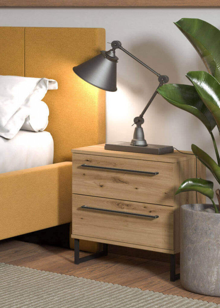 Comodino Sardinia in rovere chiaro ambientato accanto a un letto giallo senape con lampada in metallo.