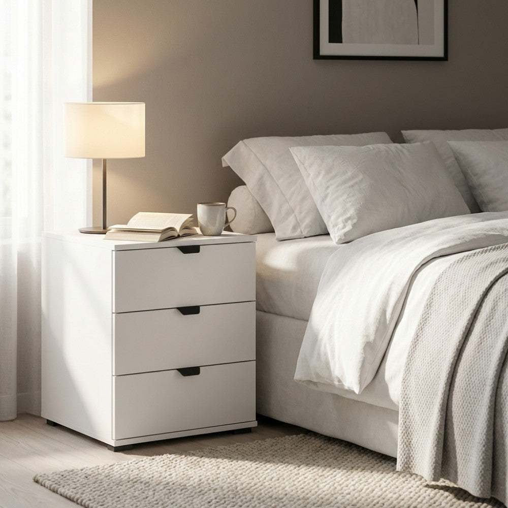 Comodino Duero bianco opaco con tre cassetti, ambientato in una camera da letto luminosa accanto a un letto matrimoniale.