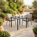 Tavolo da giardino quadrato in acciaio antracite allestito su un patio in pietra con sedie in corda intrecciata.