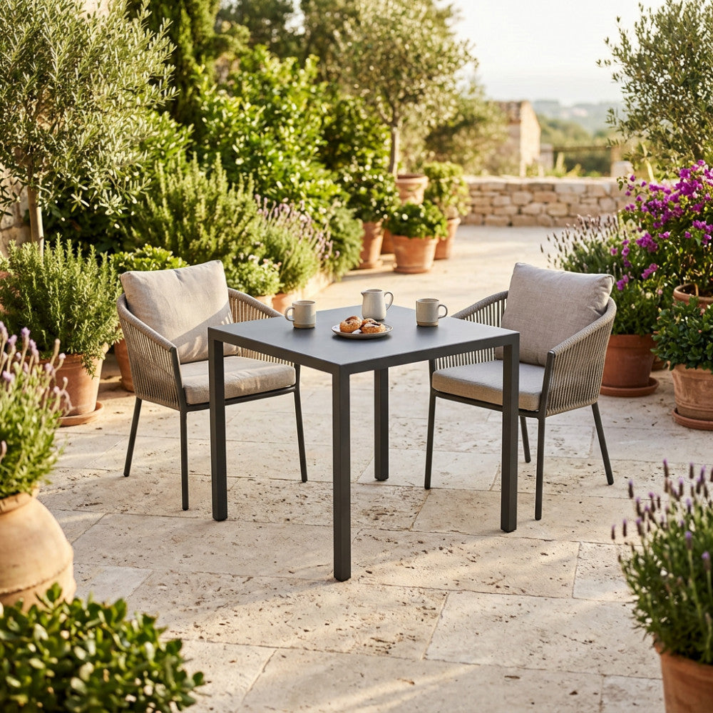 Tavolo da giardino quadrato in acciaio antracite allestito su un patio in pietra con sedie in corda intrecciata.