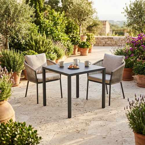 Tavolo da giardino quadrato in acciaio antracite allestito su un patio in pietra con sedie in corda intrecciata.