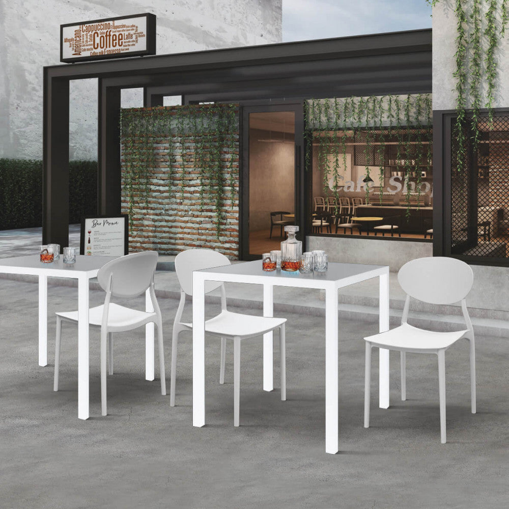 Allestimento contract con due tavoli bianchi in acciaio e sedute di design di fronte a un caffè moderno.