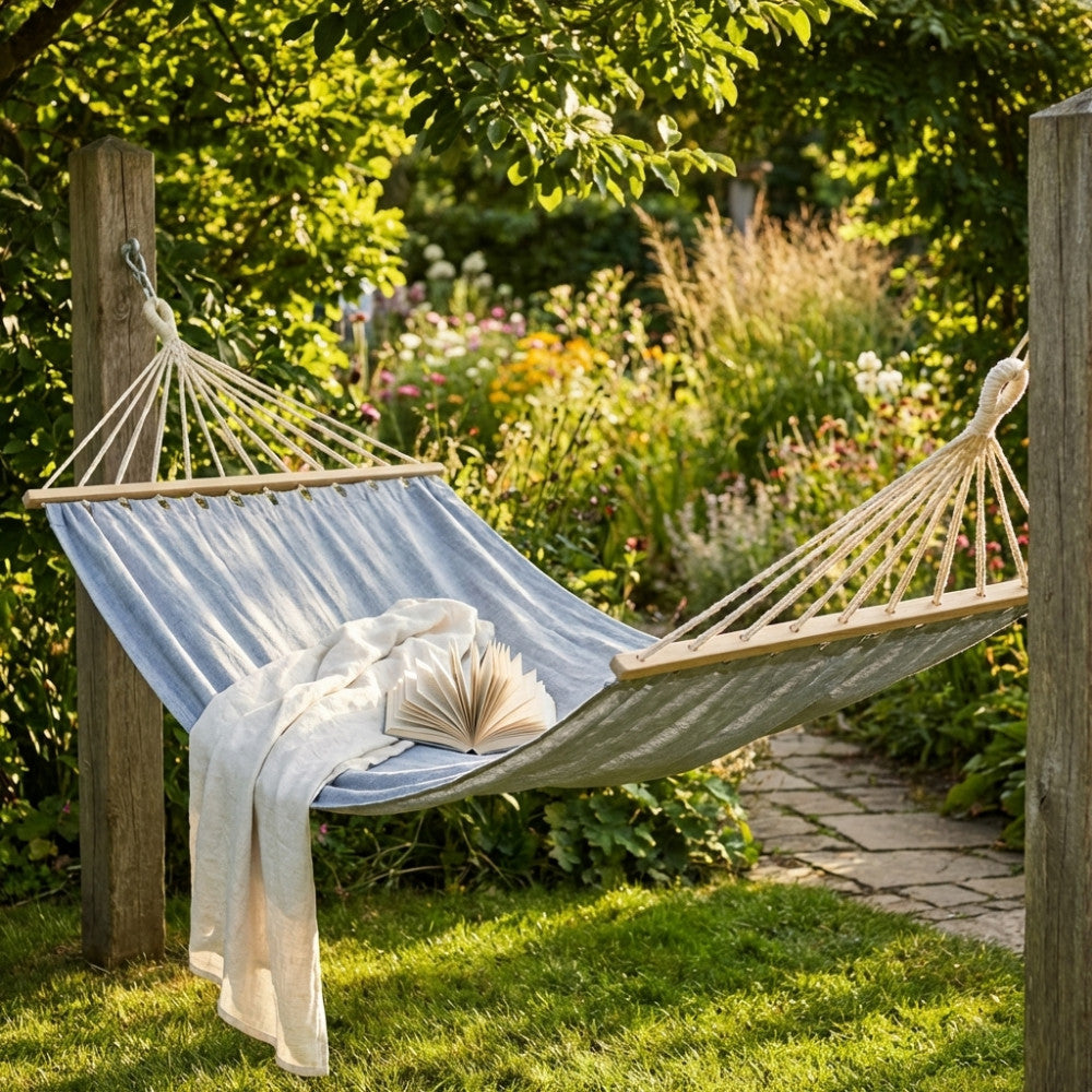 Amaca in tessuto grigio chiaro sospesa tra due pali di legno in un giardino rigoglioso e soleggiato, allestita con una coperta bianca e un libro aperto.