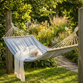 Amaca in tessuto grigio chiaro sospesa tra due pali di legno in un giardino rigoglioso e soleggiato, allestita con una coperta bianca e un libro aperto.