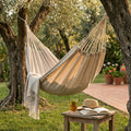Amaca Flexy in tessuto beige sospesa tra due ulivi in un giardino soleggiato, accompagnata da una coperta con frange e un tavolino in legno con bibita.