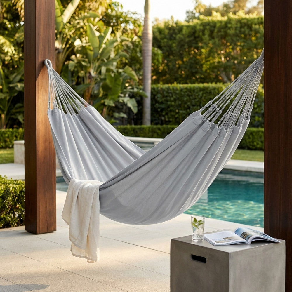 Amaca di lusso grigio chiaro installata su pali in legno a bordo piscina, con un asciugamano bianco e un tavolino in cemento.