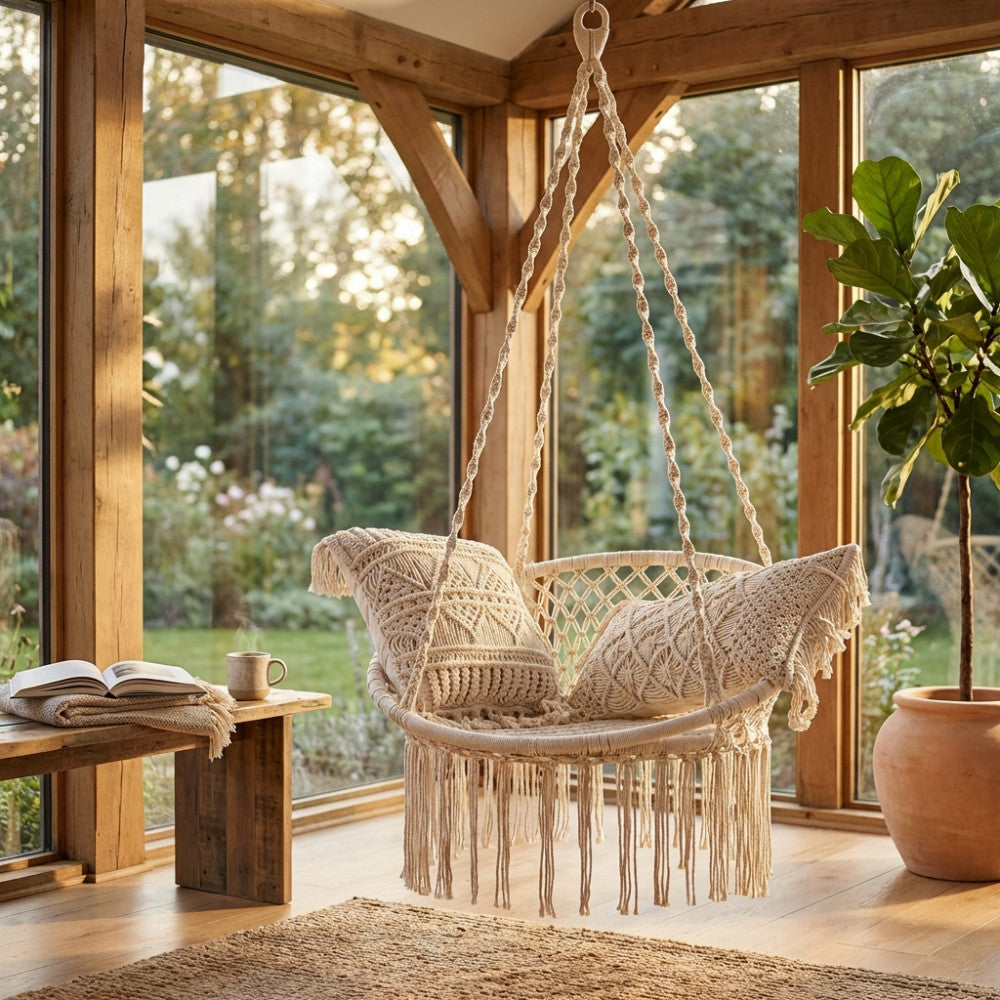 Amaca sospesa Smara in cotone ecrù con frange macramè, ambientata in un luminoso giardino d'inverno con travi in legno.