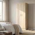 Armadio a due ante Leopold in finitura quercia chiaro, ambientato in una luminosa camera da letto moderna accanto a un letto con coperte bianche.
