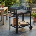 Barbecue a carbonella in metallo nero posizionato in un giardino lussuoso, con tagliere in legno, rosmarino e pinze sul ripiano laterale.