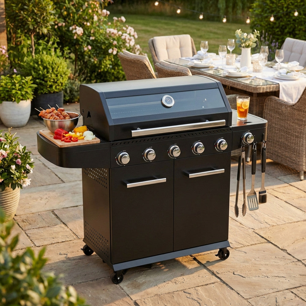 Barbecue a gas Maine 5 fuochi in metallo nero, allestito in un giardino lussuoso con tavola imbandita sullo sfondo.
