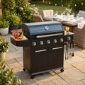 Barbecue a gas Maine 5 fuochi in metallo nero, allestito in un giardino lussuoso con tavola imbandita sullo sfondo.