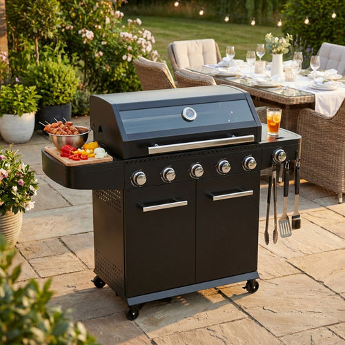 Barbecue a gas Maine 5 fuochi in metallo nero, allestito in un giardino lussuoso con tavola imbandita sullo sfondo.
