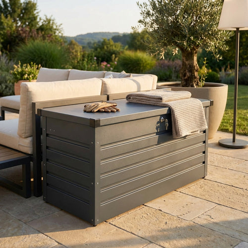 Baule da esterno New Jersey 130 in alluminio antracite, ambientato in un giardino moderno accanto a un divano lounge con coperta e guanti appoggiati sul coperchio.