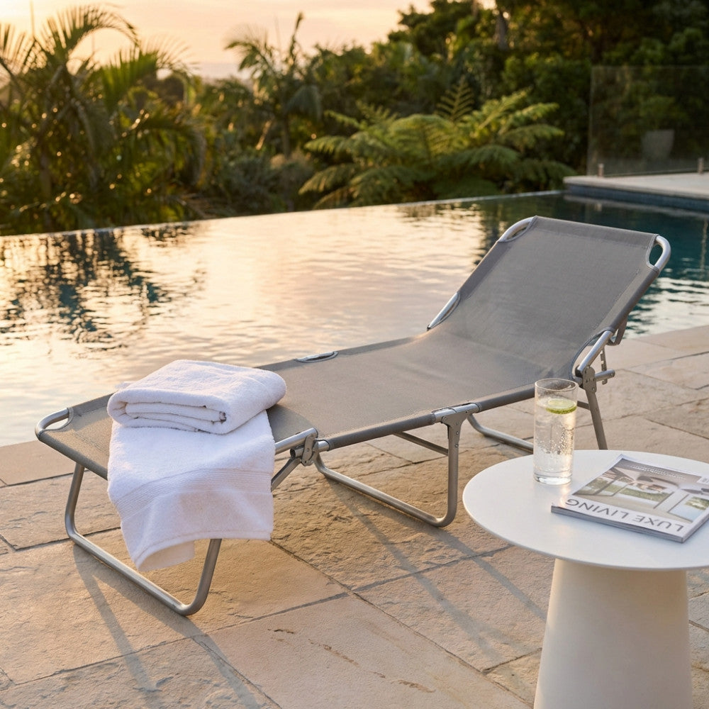 Lettino prendisole dal design essenziale immerso nella luce del tramonto, con un drink rinfrescante e una rivista su un tavolino bianco.