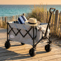 Carrello INDIAN in poliestere grigio chiaro e metallo, posizionato su una passerella in legno vicino alla spiaggia, contenente un asciugamano a righe e un cappello di paglia.