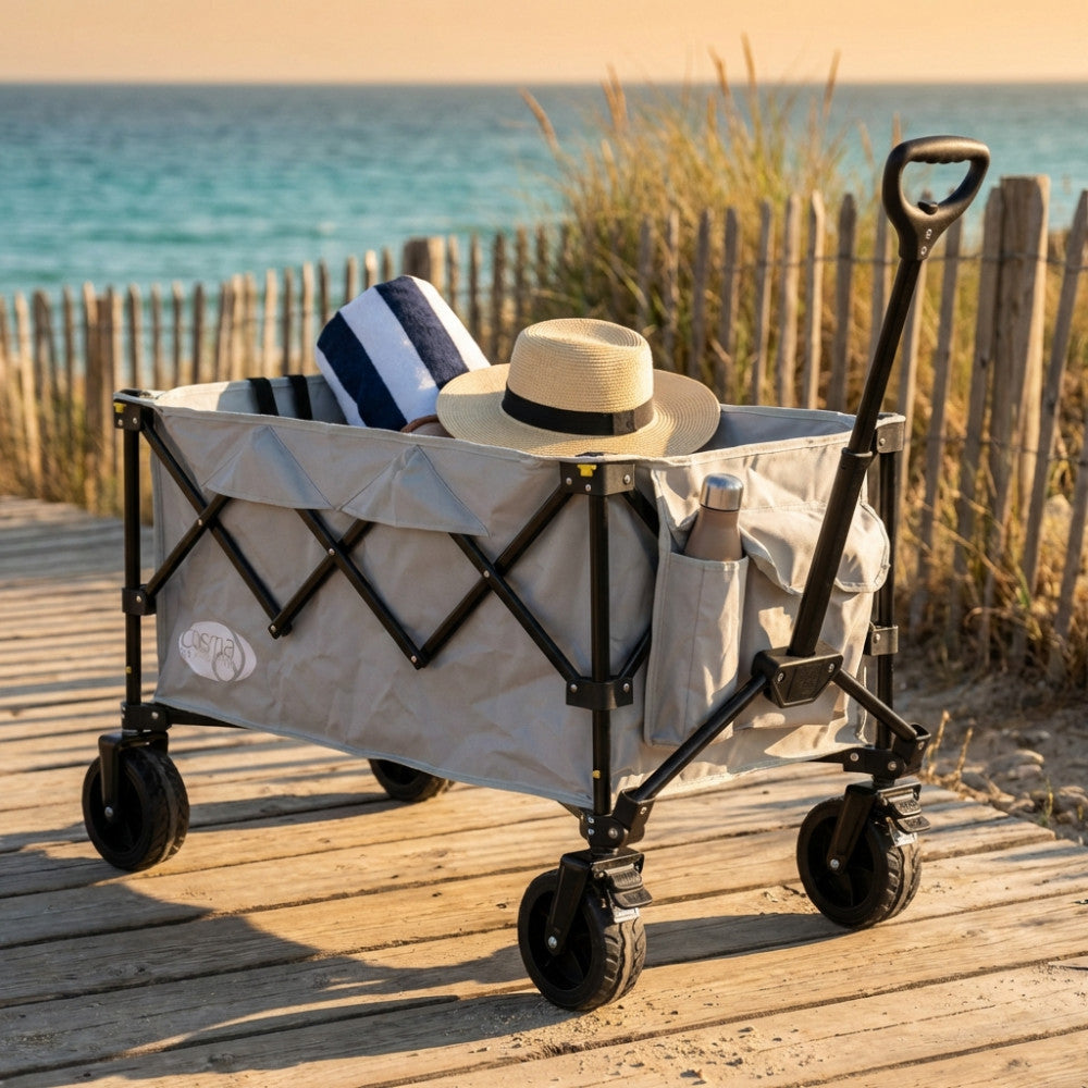 Carrello INDIAN in poliestere grigio chiaro e metallo, posizionato su una passerella in legno vicino alla spiaggia, contenente un asciugamano a righe e un cappello di paglia.