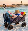 Carrello Pacific blu in acciaio, caricato con teli mare e accessori, posizionato sulla sabbia al tramonto.