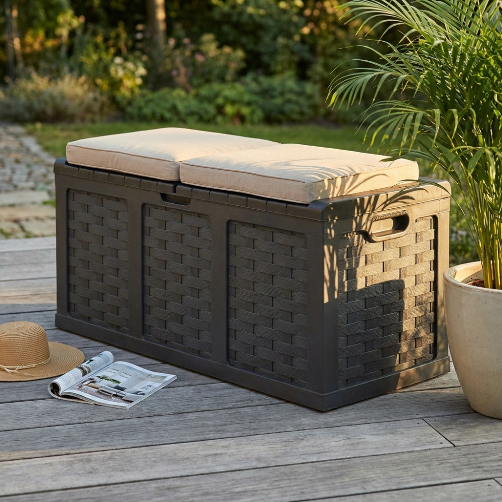 Cassapanca da esterno marrone effetto rattan posizionata su un decking in legno, allestita con cuscini chiari e affiancata da una pianta verde e una rivista.