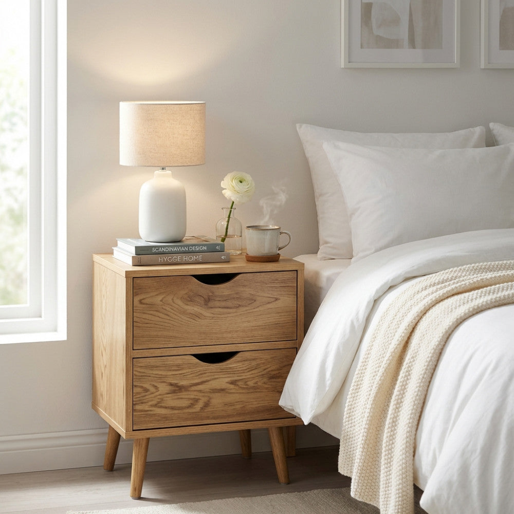 Vista frontale del comodino in legno moderno accanto a un letto con lampada in ceramica bianca e fiori.