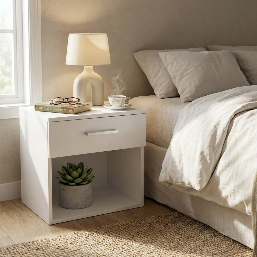 Comodino bianco in truciolare bilaminato posizionato accanto a un letto, decorato con una lampada accesa e una tazza fumante.