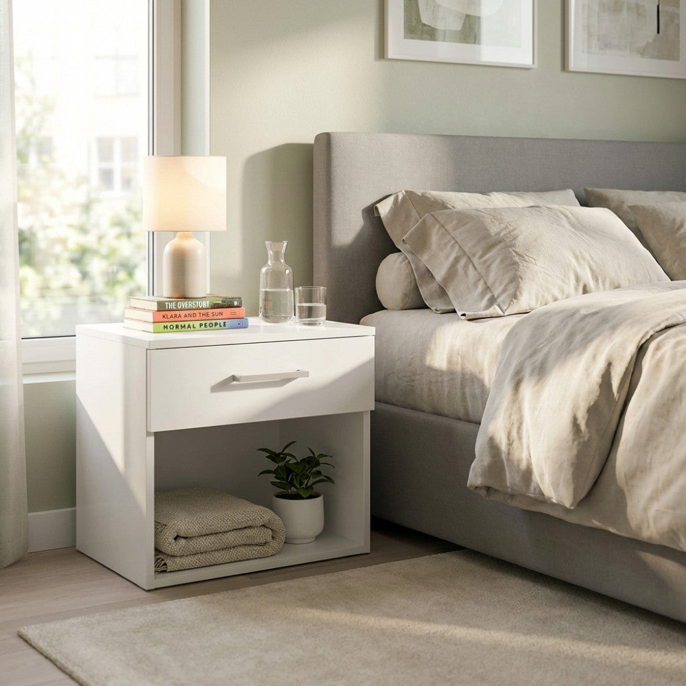 Elegante comodino bianco con cassetto superiore e vano a giorno, ambientato in una luminosa camera da letto contemporanea.