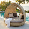 Conchiglione Palma in wicker marrone posizionato a bordo piscina con cuscini beige e marroni, asciugamano bianco e libro aperto.