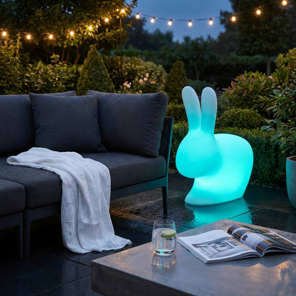 Coniglietto luminoso Volans in polietilene azzurro posizionato su un patio esterno accanto a un divano scuro.
