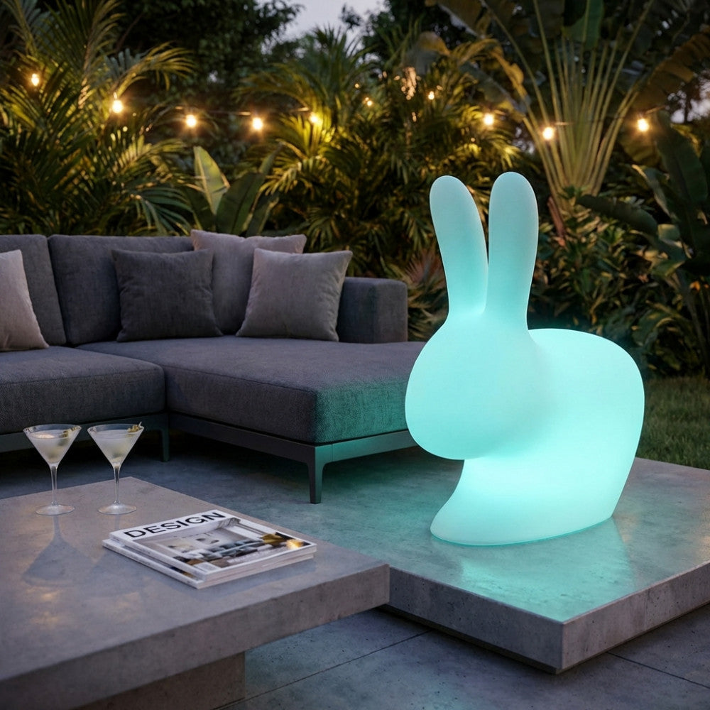 Lampada moderna Volans a forma di coniglio illuminata di azzurro in un giardino tropicale con tavolino in cemento.