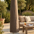 Copertura per ombrellone Giove in poliestere taupe, posizionata su un patio in pietra accanto a un divano in legno con cuscini beige e un tavolino da caffè.