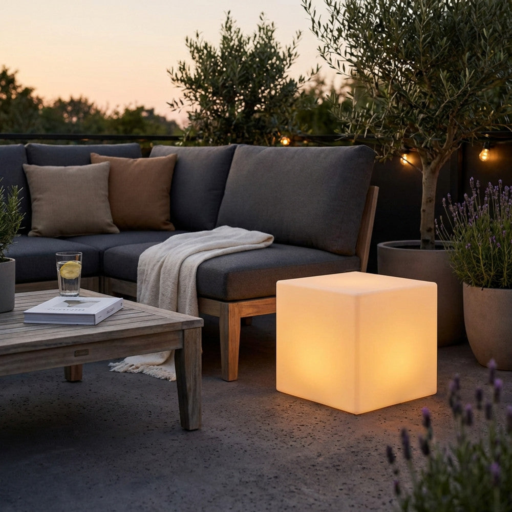 Cubo luminoso in polipropilene bianco che illumina un patio esterno accanto a un divano grigio scuro al tramonto.