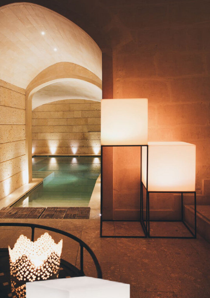 Due cubi luminosi elegantemente impilati su strutture in metallo nero accanto a una piscina coperta con archi in pietra.