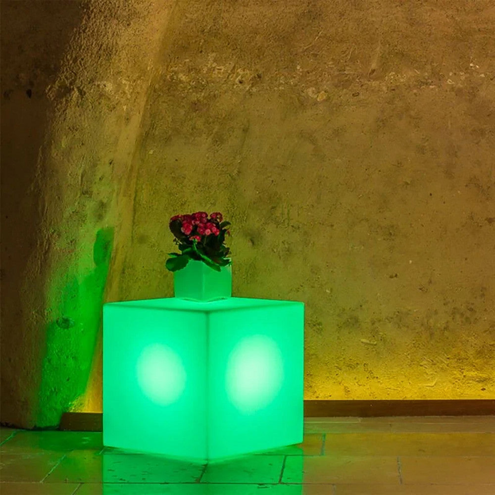 Cubo luminoso illuminato di verde vibrante, utilizzato come piedistallo per una piccola pianta in vaso in un interno rustico.