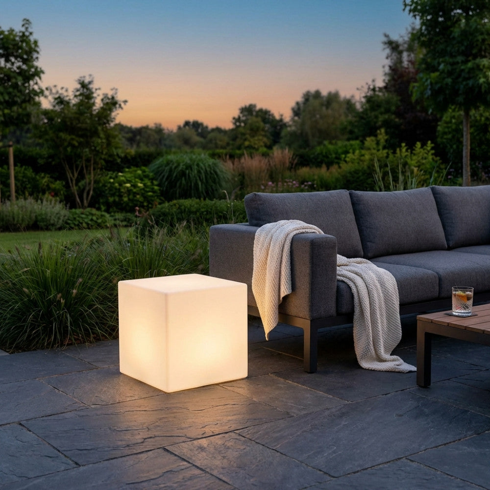 Illuminazione geometrica a forma di cubo accesa al tramonto su una pavimentazione in ardesia, vicino a un divano da giardino scuro.