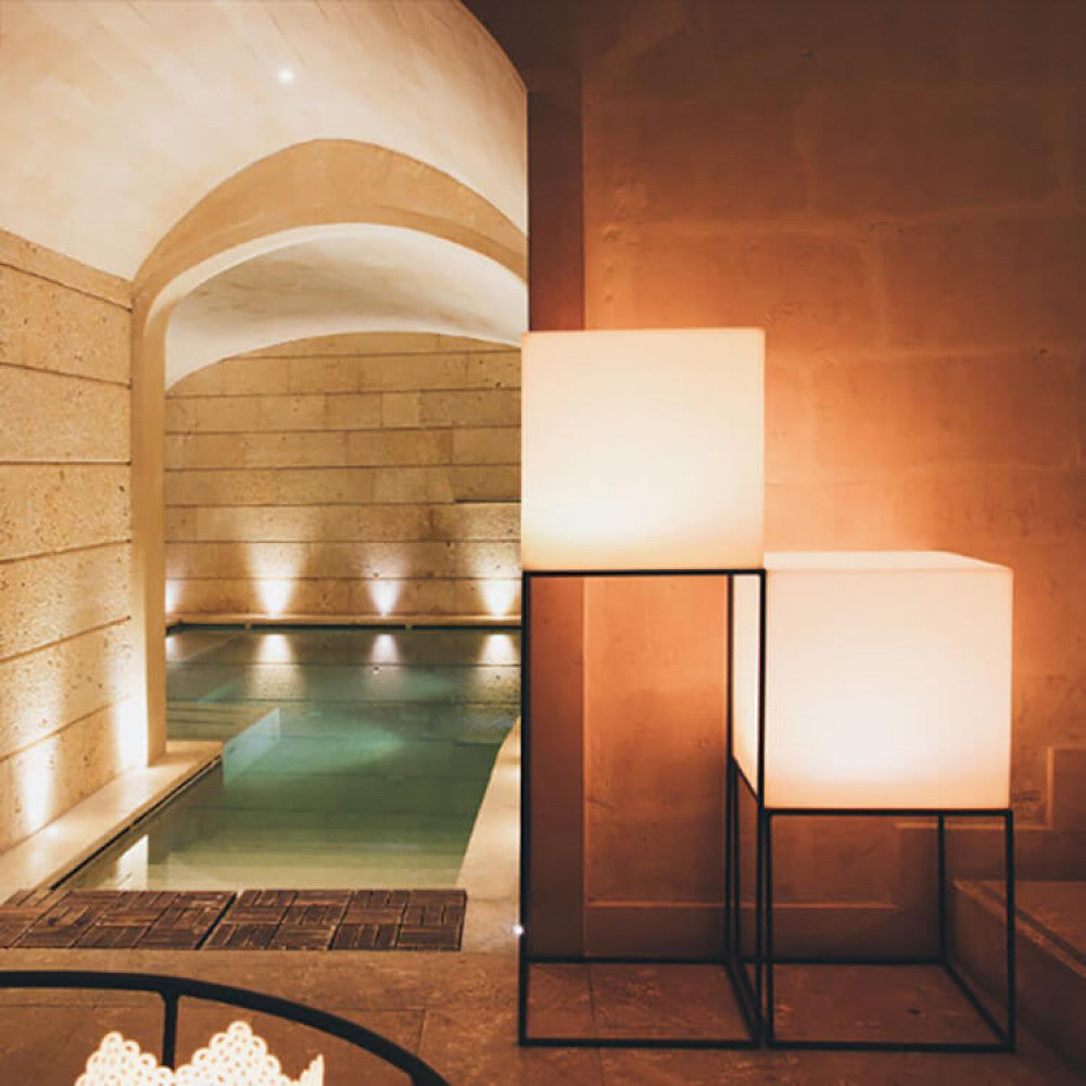Due lampade a cubo luminoso installate su eleganti piedistalli in metallo nero all'interno di una lussuosa area piscina coperta in pietra.