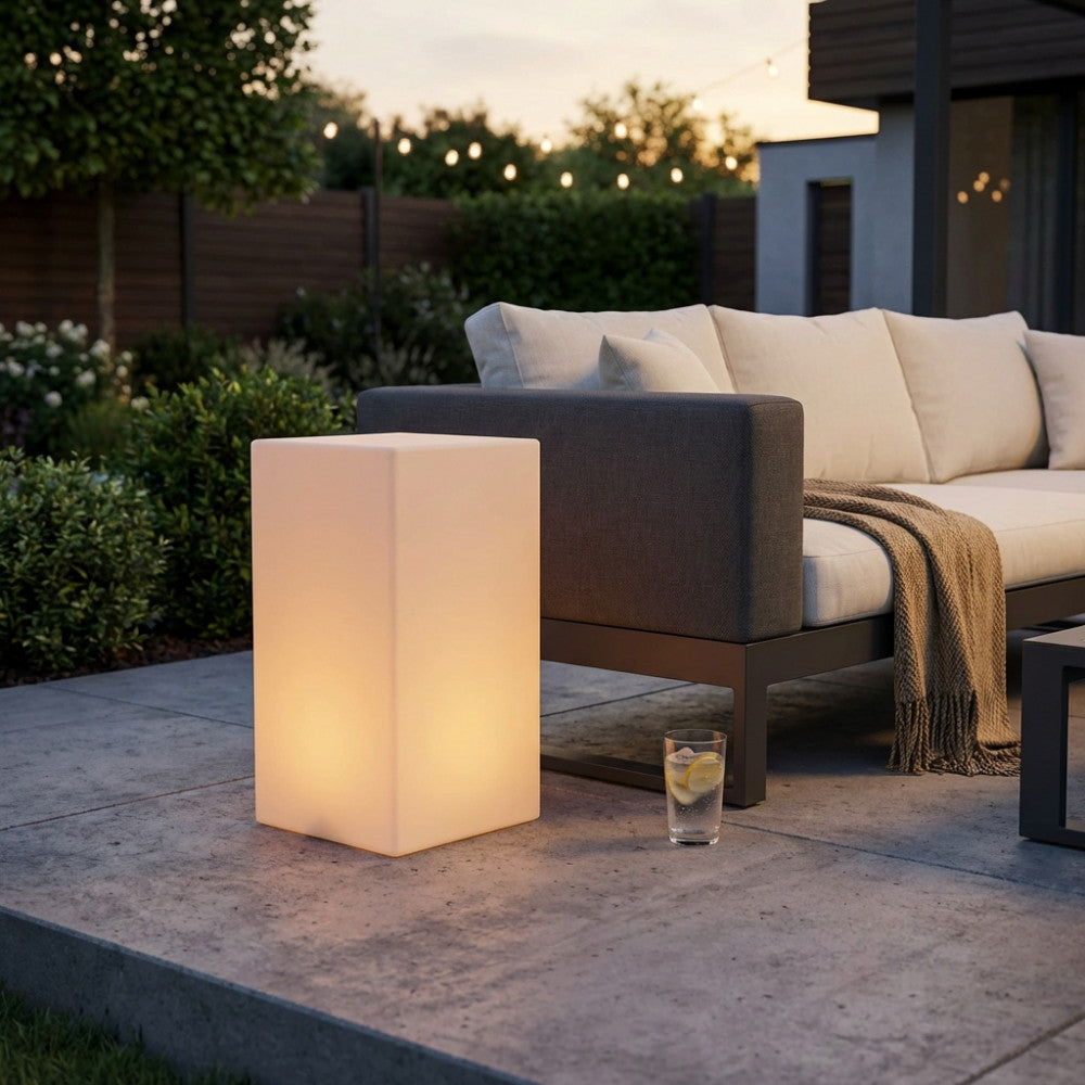 Lampada da terra Cubo Indus illuminata con luce calda accanto a un divano da esterni.
