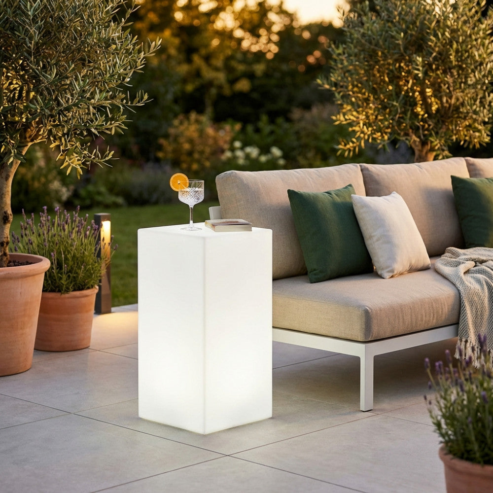 Cubo luminoso bianco utilizzato come tavolino da esterno accanto a un divano in tessuto, con un cocktail e un libro, in un giardino al tramonto.