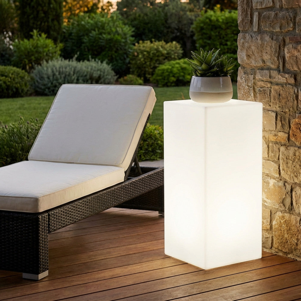 Cubo luminoso in polipropilene posizionato su un ponte in legno accanto a un lettino prendisole scuro, decorato con una pianta in vaso.