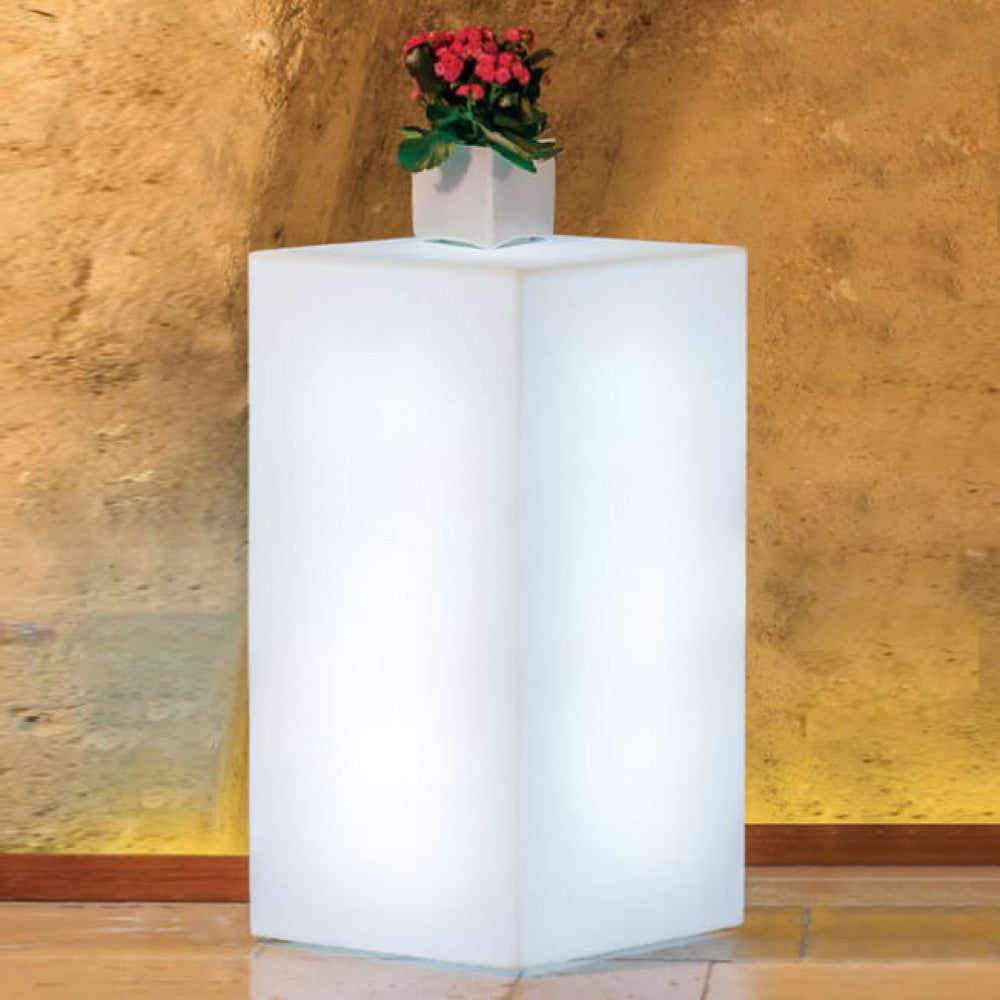 Cubo luminoso acceso in un ambiente interno con pareti in pietra rustica, sormontato da un vaso con fiori rossi.