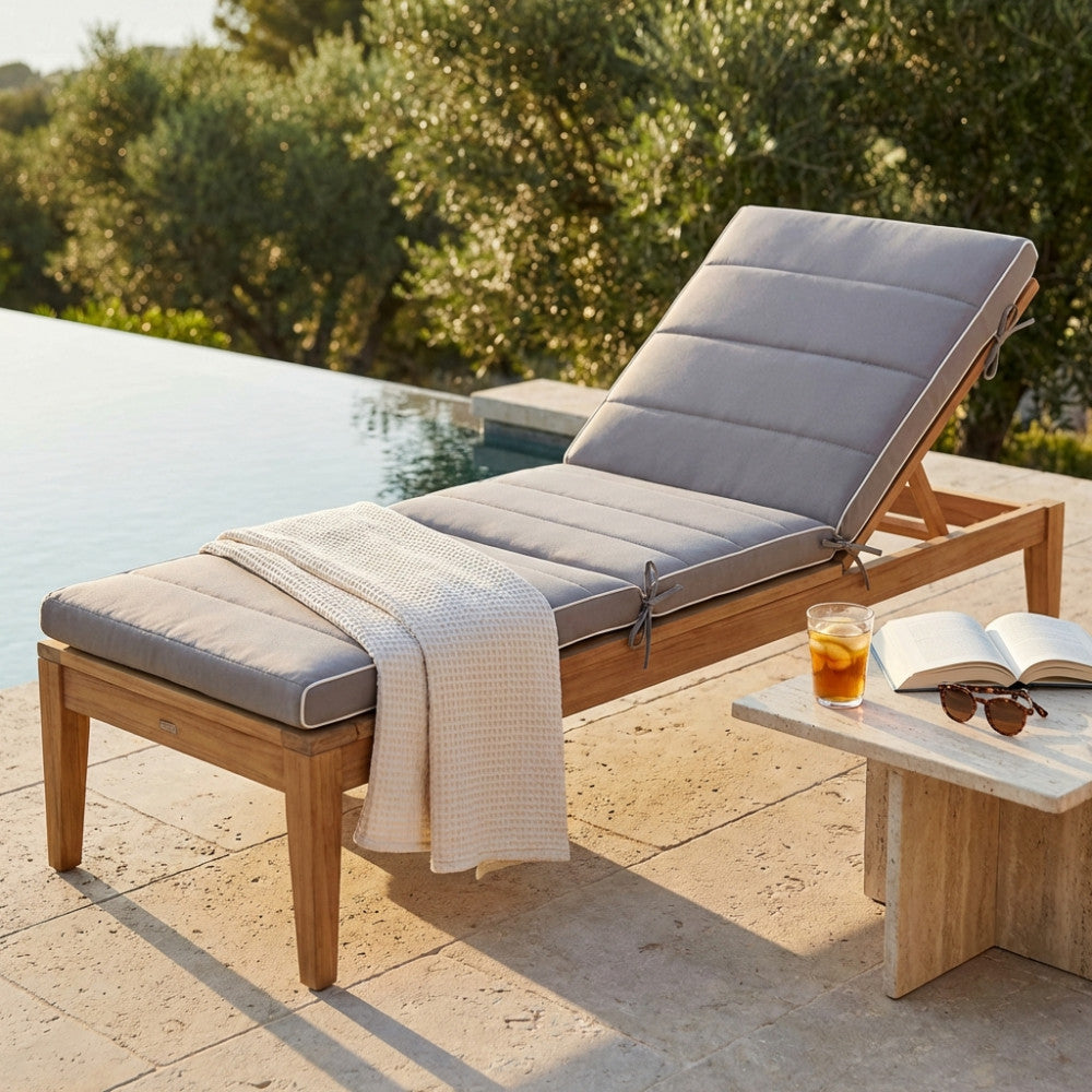 Cuscino per lettino King grigio posizionato su una sdraio in legno di teak a bordo piscina, allestito con una coperta bianca testurizzata e un bicchiere di tè freddo.