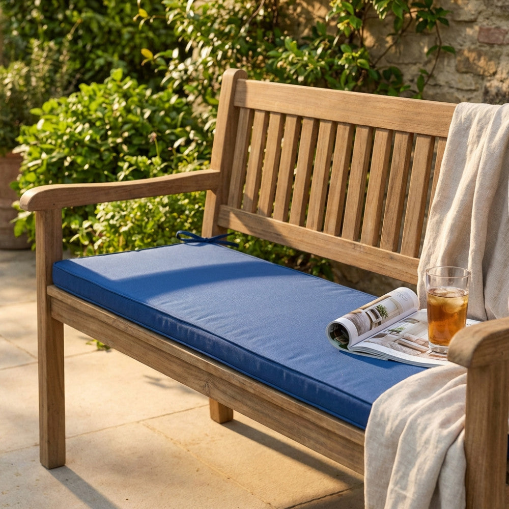 Cuscino Real 110 blu scuro posizionato su una panchina in legno di teak in un giardino soleggiato, affiancato da una coperta chiara e un bicchiere.