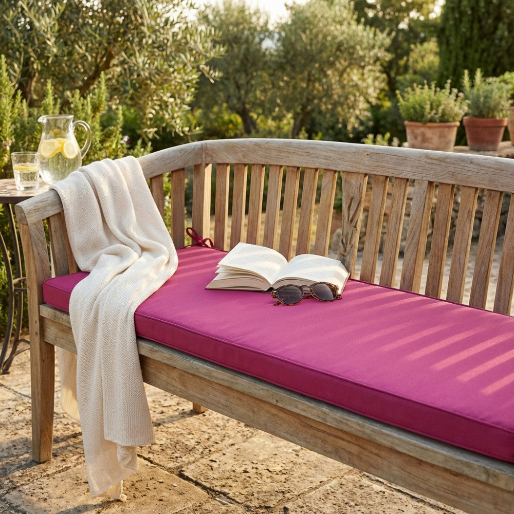 Cuscino magenta Real 110 posizionato su una panca in legno da esterni, allestito con un libro aperto e occhiali da sole.