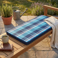 Cuscino Real 110 Quadri Blu posizionato su una panca in legno in un giardino soleggiato, con una tazza e un libro accanto.
