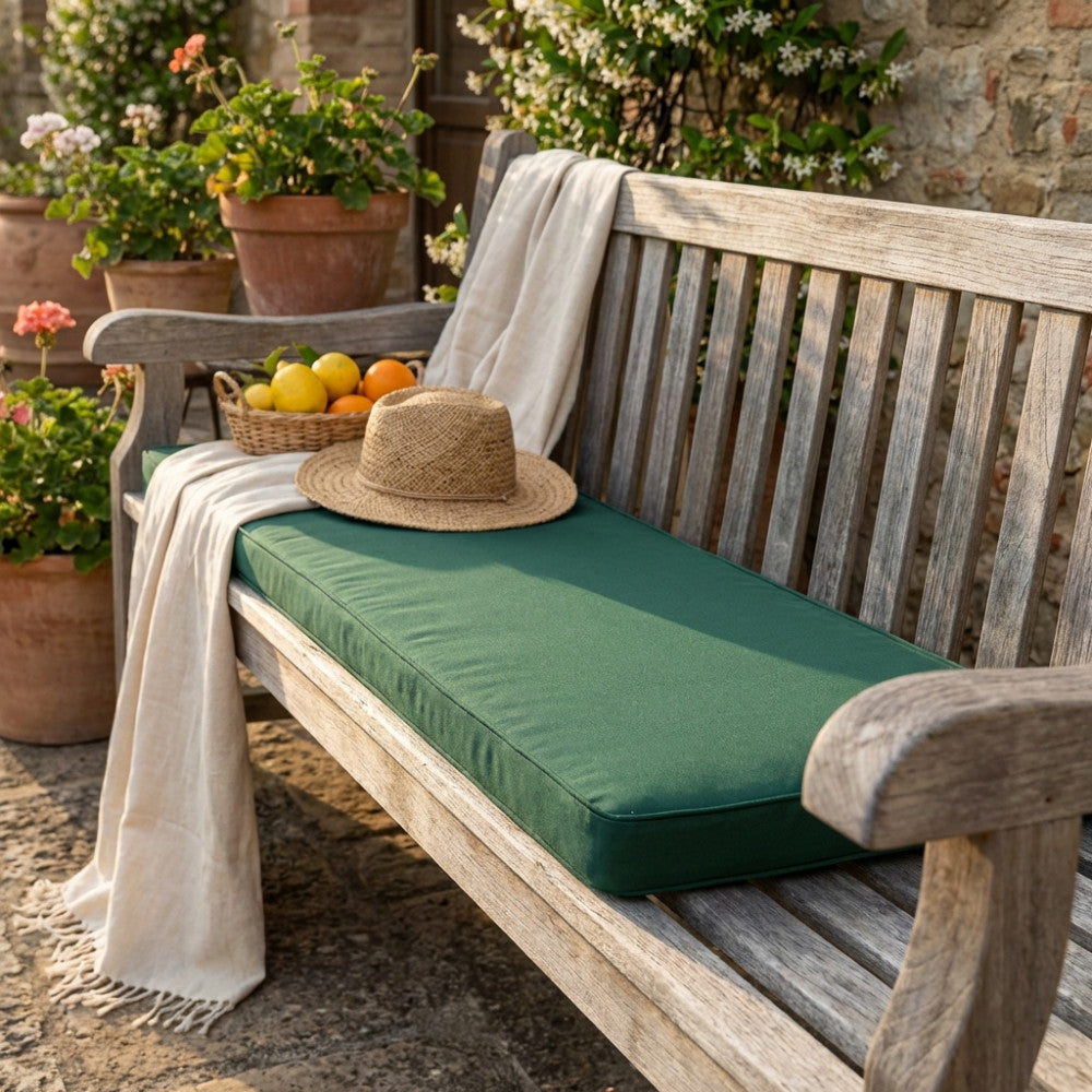 Cuscino Real 110 verde scuro posizionato su una panca in legno rustico in un giardino soleggiato, decorato con un cappello di paglia e un cesto di agrumi.