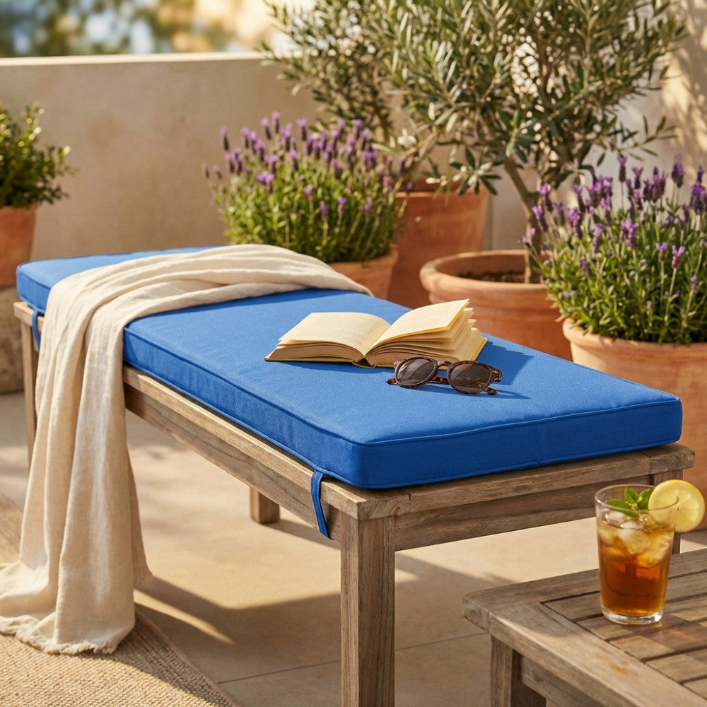 Cuscino rettangolare blu scuro posizionato su una panca in legno senza schienale, allestita con un libro aperto, occhiali da sole e una bevanda fresca in un patio soleggiato.