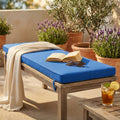 Cuscino rettangolare blu scuro posizionato su una panca in legno senza schienale, allestita con un libro aperto, occhiali da sole e una bevanda fresca in un patio soleggiato.