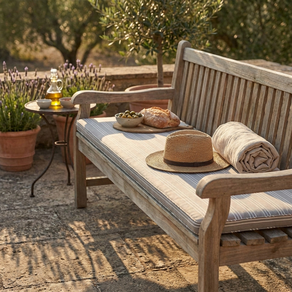 Dettaglio del cuscino moderno su panca in legno rustico, allestito con cappello di paglia, pane fresco e olive in un giardino.
