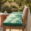 Dettaglio del cuscino Real 150 verde scuro posizionato su una panca in legno in un giardino soleggiato, affiancato da un tavolino in terracotta con tazza e libro.