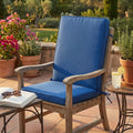 Cuscino Real Medio Blu Scuro posizionato su una sedia in legno da esterni, accanto a un tavolino con una bevanda fresca, in un giardino soleggiato.