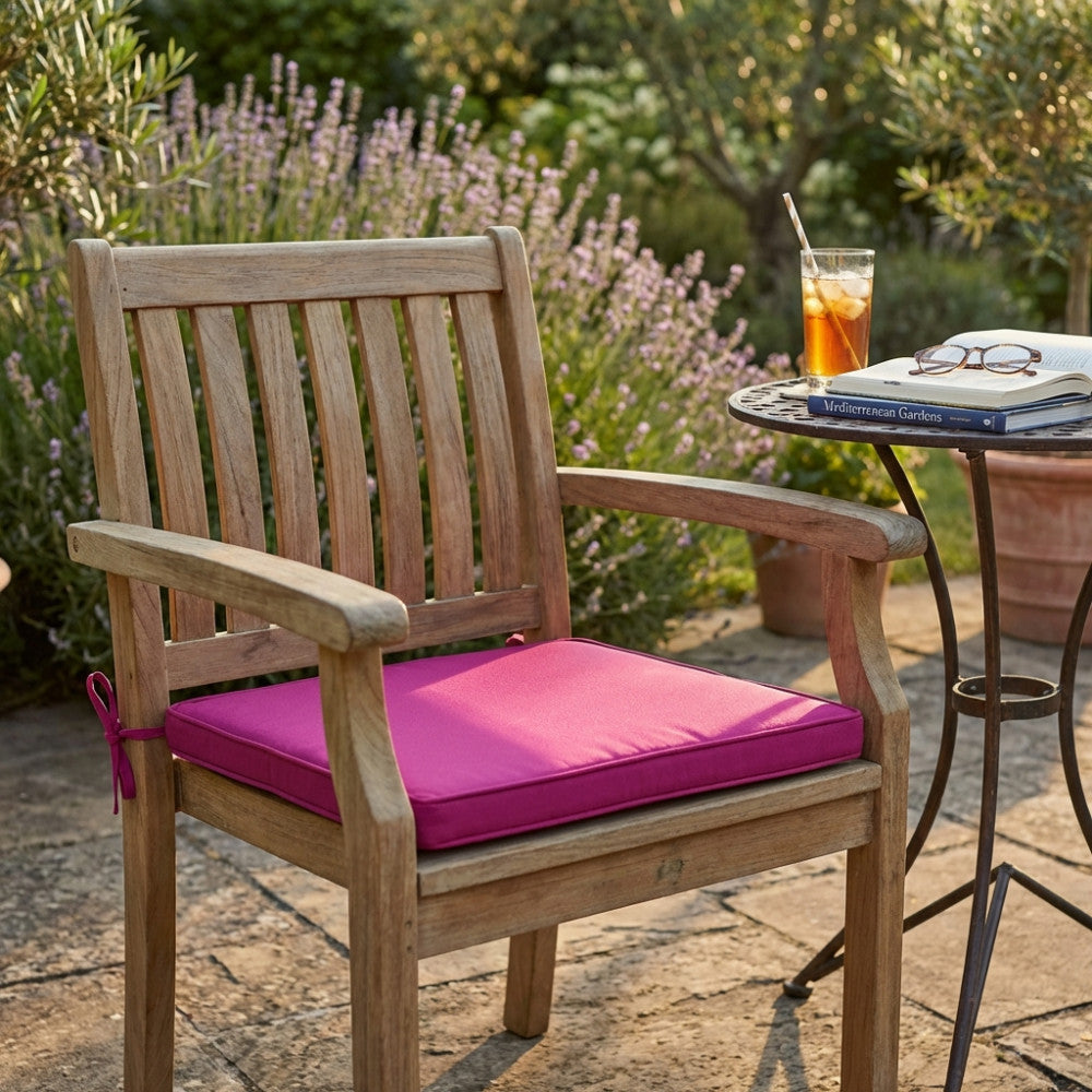 Sedia in legno di teak con cuscino magenta Real Seduta in un elegante patio esterno, accanto a un tavolino con libro e bibita rinfrescante.