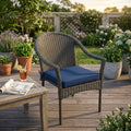 Cuscino Real Seduta Large blu scuro posizionato su una poltroncina in rattan intrecciato in un giardino elegante.